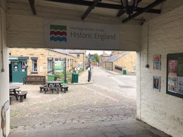 Archway into the heritage centre with an Historic England sign on the roof. Picnic benches and stone buildings can be seen through the rectangular arch.