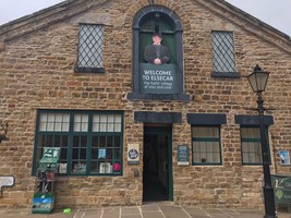 Visitor Centre entrance. An open green door below a sign of a man with a top hat saying 'Welcome to Elsecar Heritage Centre'. Windows with green frames and an old Victorian street lamp.