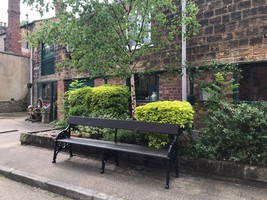 A brown bench on a pavement with bushes behind it