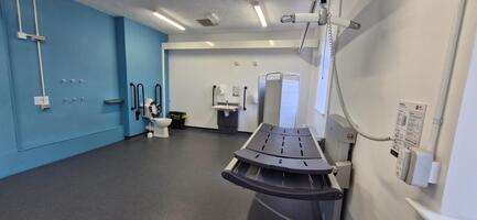 An accessible restroom at Elsecar Heritage Centre features a changing table, support bars, a toilet, sink, shower area, blue and white walls, and various accessibility aids.