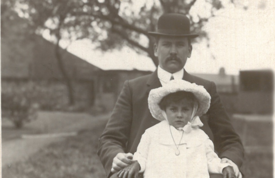 A man in a suit and bowler hat sits outdoors with a young child dressed in light clothing and a wide-brimmed hat on his lap. The background is blurred, showing trees and buildings. The photo is in black and white.