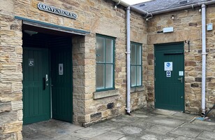 A stone building at Elsecar Heritage Centre features three green doors labelled as public restrooms, including one with an accessibility symbol and one with a Changing Places sign. The ground is paved with large stone slabs.