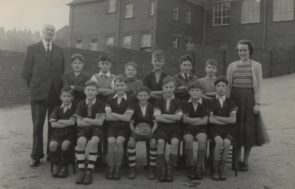 A black-and-white photo of a boys' soccer team from the mid-20th century, with 13 boys in uniform, one holding a ball labeled "1949-50," posing outdoors with a man in a suit and a woman in a sweater and skirt.