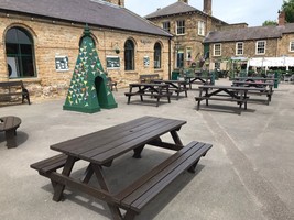 Brown rectangular picnic benches in an open space. A green triangular structure with bird images stuck onto it is a piece of public art to the left of the image.